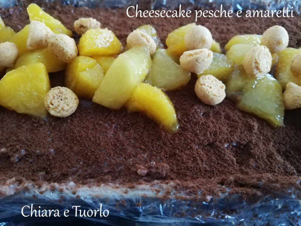 particolare della Cheesecake pesche e amaretti vista dall'alto, spolverata di cacao e cosparsa al centro da pezzetti di pesca e mini amaretti