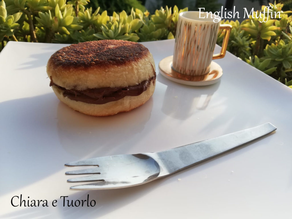 Un english muffin farcito di Nutella su un piatto bianco con posata e tazzina da caffe' dorata